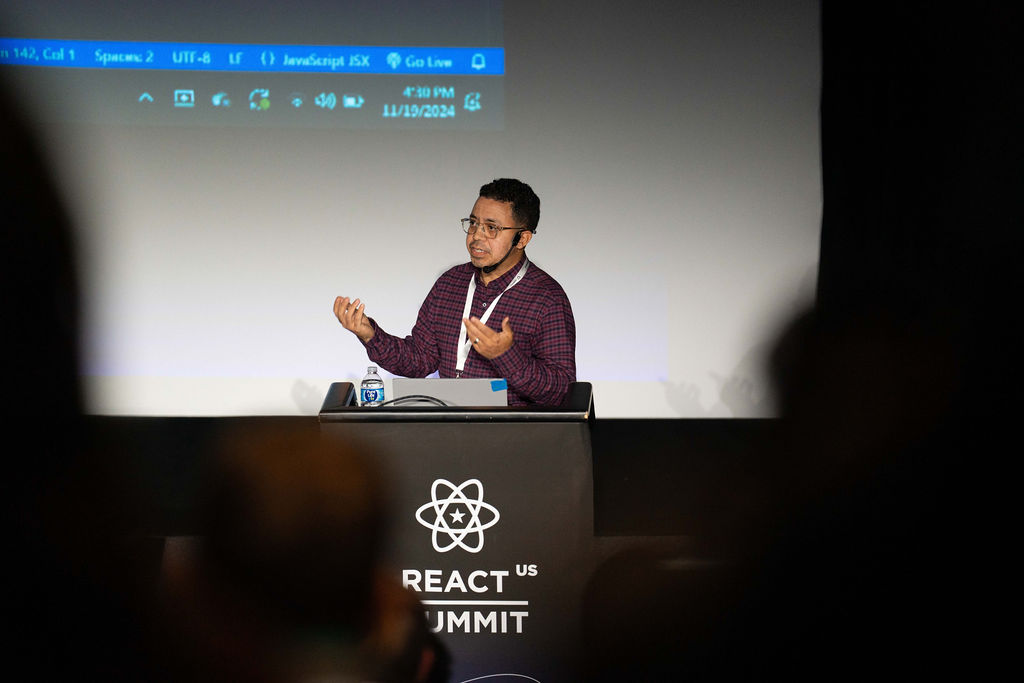 Tony Alicea standing at a podium which says 'React Summit' on the front giving a technical talk on React and JavaScript.